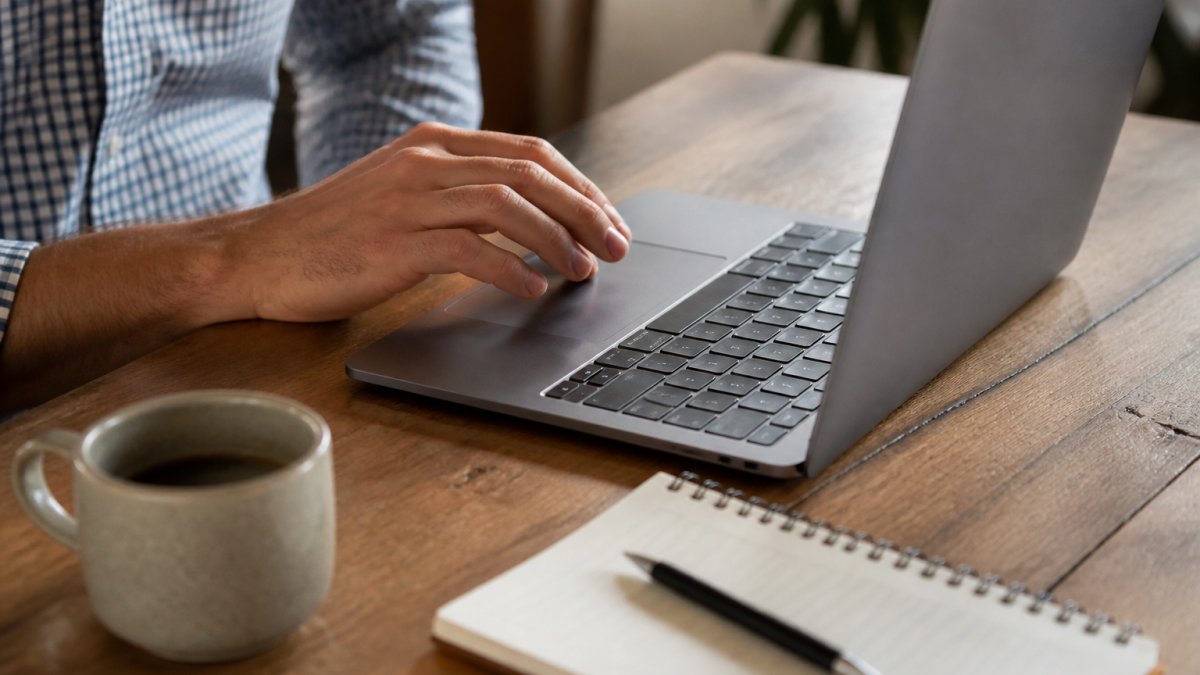 Nahaufnahme einer Hand, die auf einem Laptop-Touchpad ein Kontaktformular mit nur drei Feldern ausfüllt, mit einer Tasse Kaffee und Notizbuch daneben auf dem Schreibtisch